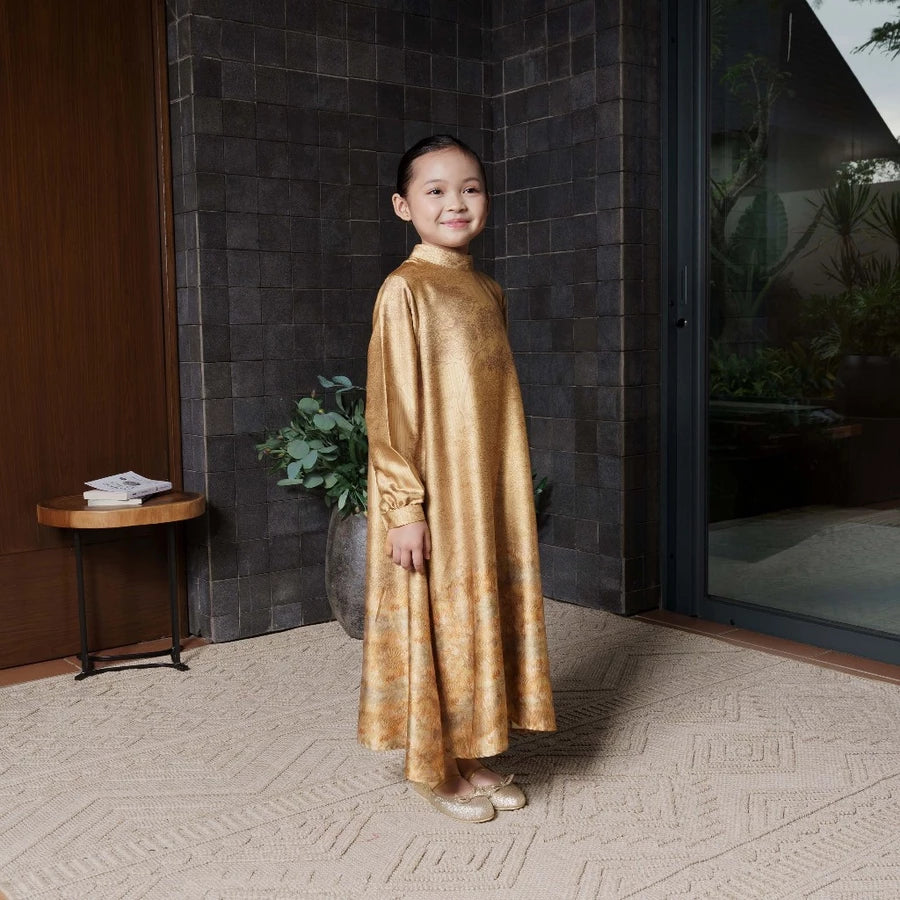 Child wearing a gold dress by Jenna & Kaia standing in a room with a textured wall and a small table.