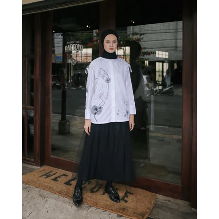 A woman wearing a white blouse by Maima with long sleeves and a round neckline, paired with a maxi skirt. The blouse features a floral pattern and she is standing in front of a glass door.