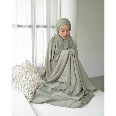 Woman wearing a light green mukena by Mistyvori sitting on a white surface with a white curtain in the background
