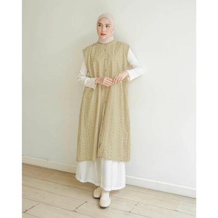 Woman wearing a butter-colored long vest by Mistyvori over a white outfit against a plain white wall.