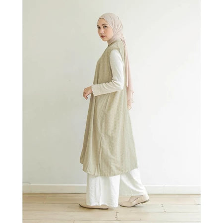Woman wearing a khaki long vest by Mistyvori over a white shirt with a hijab, standing against a plain white wall.