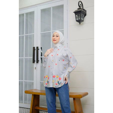 A woman wearing a grey long-sleeved shirt by Abame with a floral pattern, standing on a bench with her hand raised to her face, in front of a door with glass panels and a wall-mounted lamp.