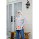 A woman wearing a grey long-sleeved shirt by Abame with a floral pattern, standing on a bench with her hand raised to her face, in front of a door with glass panels and a wall-mounted lamp.