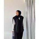 Person wearing a black hijab and black vest by Abame against a white wall with a curtain.