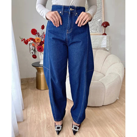 A woman standing in an indoor setting wearing dark blue barrel jeans and white top, with decorative flowers and furniture in the background.

Seorang wanita berdiri di dalam ruangan, mengenakan celana denim model barrel berwarna biru tua dan atasan putih, dengan hiasan bunga dan perabotan di latar belakang.