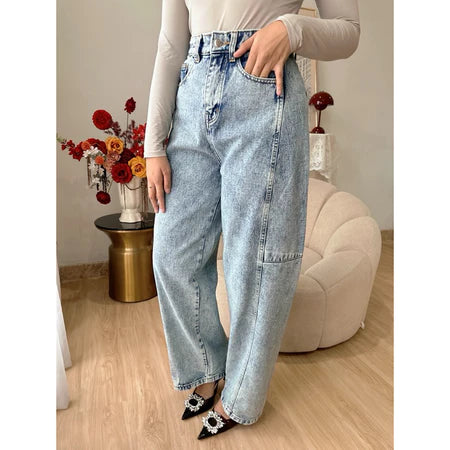 A person standing in an indoor setting wearing light blue washed barrel jeans with a high waist and straight leg from Haidee Orlin, paired with a beige top and black and white shoes.