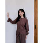 Woman wearing a brown blouse by Renaette against a white wall.