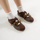 Brown flat shoes with strap bow laces by Dianable worn with white socks on a light background