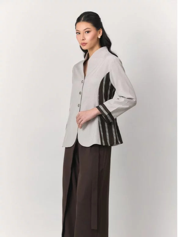 A woman modeling a light gray asymmetrical button-front blazer by Studio Tui with black and gray striped fabric panels on the sleeves and lower hem, paired with dark brown wide-leg trousers, against a plain light gray studio background.