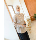 Woman wearing a beige shirt by Abame with lace details and a hijab, holding a black cup indoors.