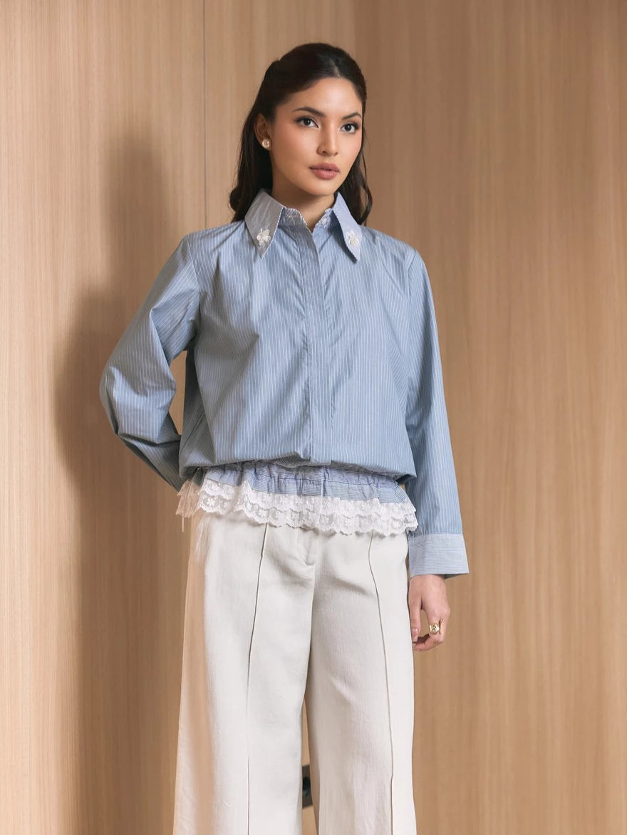 A woman models a light blue and white vertically striped button-down shirt by Studio Tui with a white lace ruffle hem, paired with cream wide-leg trousers, set against a warm wood-paneled background.