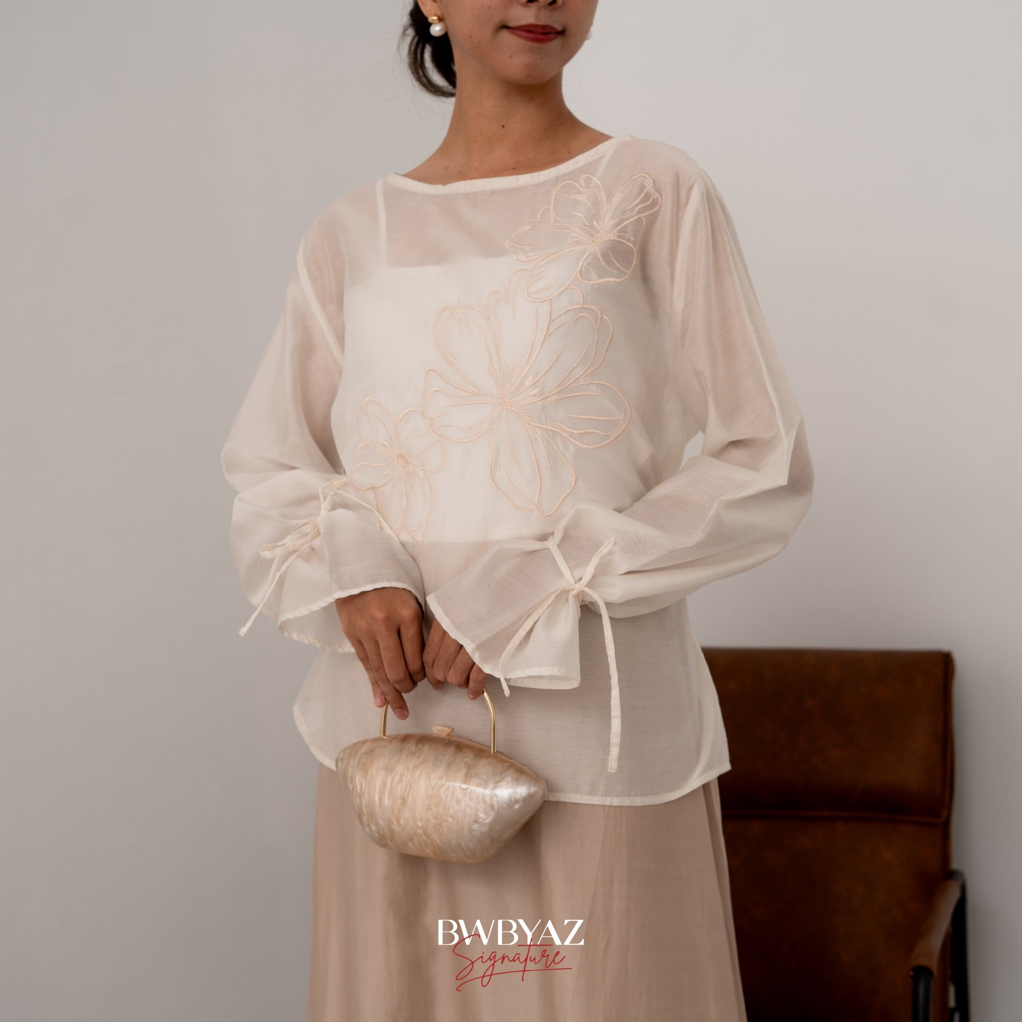 A woman wearing a white blouse with floral embroidery on the front, made from Luminous Sheer Tencel, with tie accents on the sleeves and a small loop at the waist.