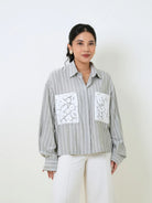 A woman wearing a light gray and white vertically striped button-up shirt by Studio Tui with two white lace pocket flaps, paired with white pants, standing against a plain off-white background.