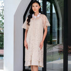 Woman wearing a beige lace outer dress by Holy Peony standing in front of a glass door.