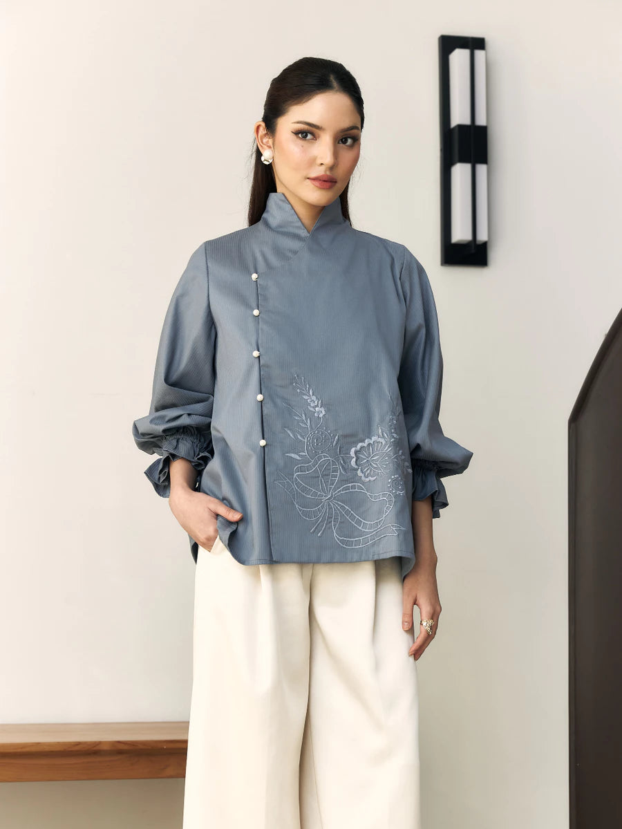 Woman wearing a blue blouse with floral embroidery by Studio Tui and white pants indoors.