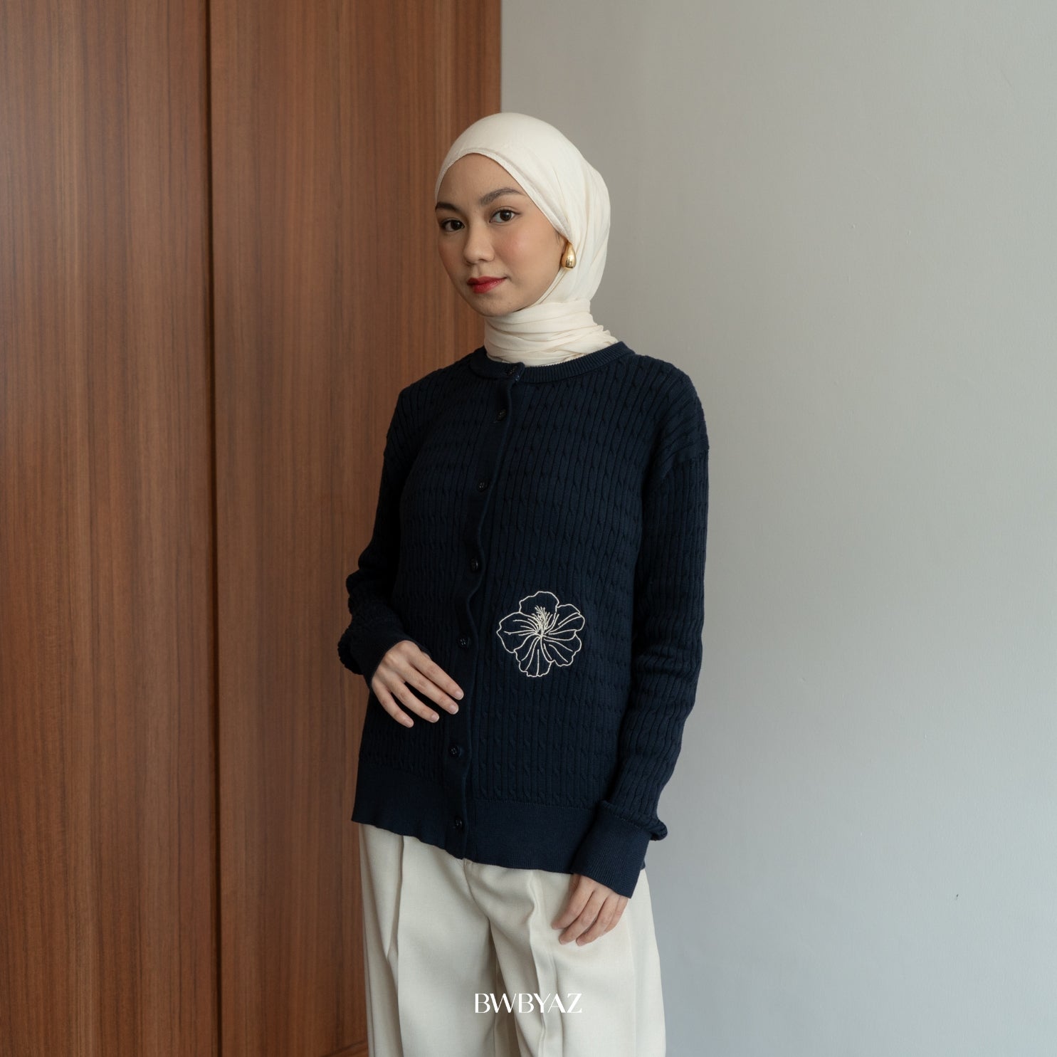 Woman wearing a navy blue cable knit long sleeve button-up cardigan with white floral embroidery on the pocket from BWBYAZ, styled modestly with a cream hijab and ivory trousers.