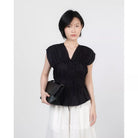 A woman modeling a black sleeveless peplum top by Kasyalia with a gathered V-neckline, paired with white pants and holding a black clutch bag, set against a plain light gray background.