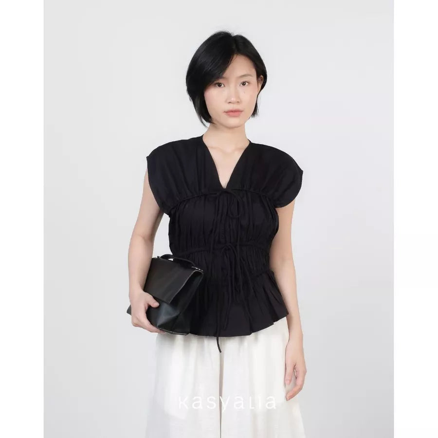 A woman modeling a black sleeveless peplum top by Kasyalia with a gathered V-neckline, paired with white pants and holding a black clutch bag, set against a plain light gray background.