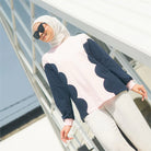 Woman wearing a pink sweater with navy blue patterns by Casa Elana and a hijab, standing on a white railing.