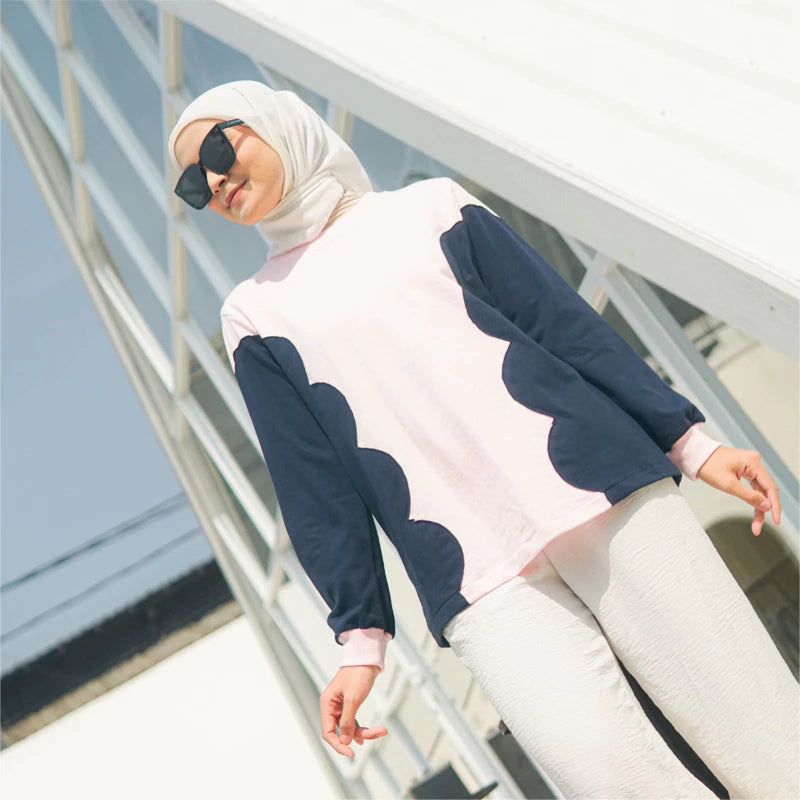 Woman wearing a pink sweater with navy blue patterns by Casa Elana and a hijab, standing on a white railing.