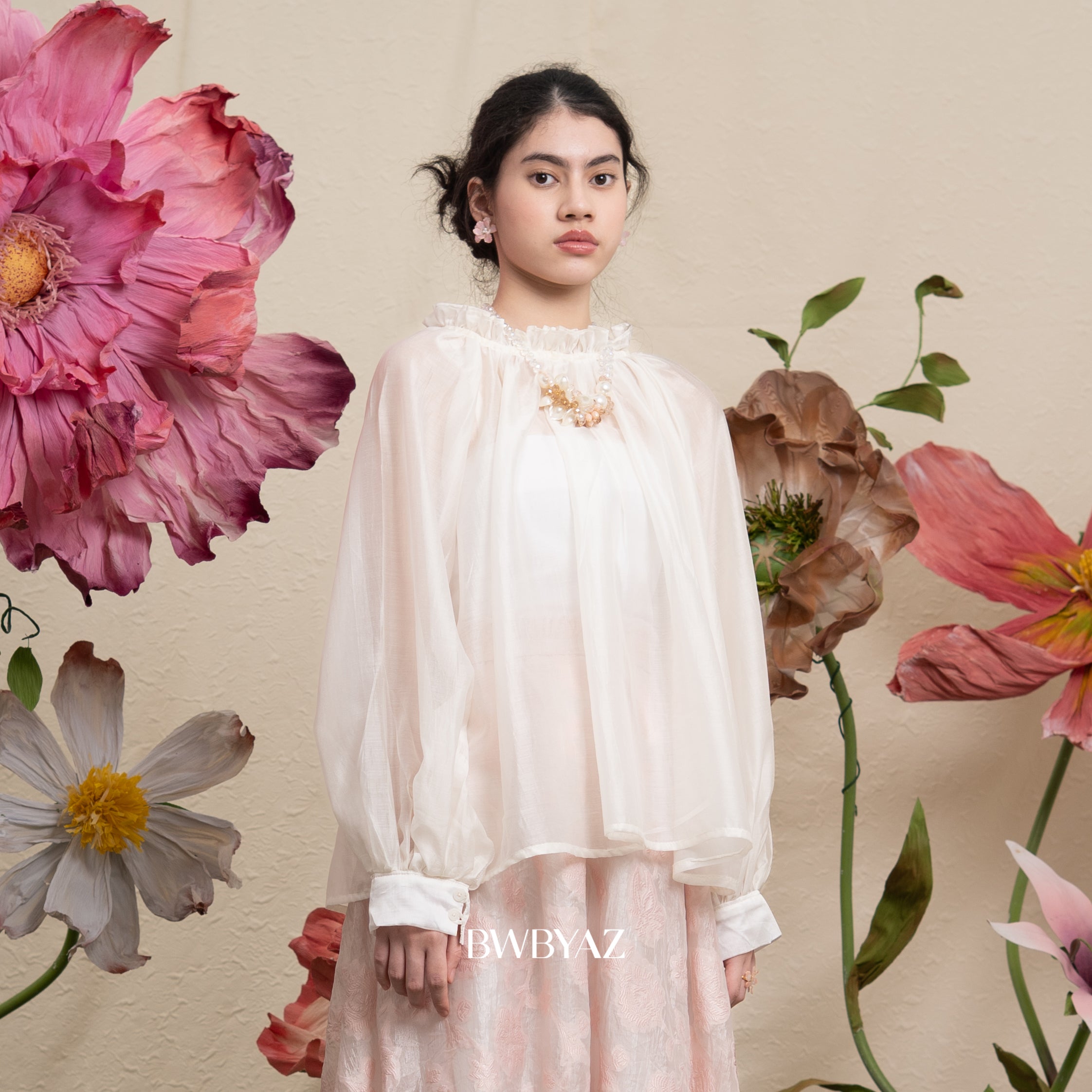 Woman in a white puff blouse standing among large flowers with 'BWBYAZ' branding.