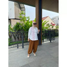 Woman in a white top by Abame and brown pants standing outdoors with greenery and buildings in the background