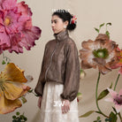 Woman in a dark brown floral-patterned lace jacket and white pants standing among large flowers with 'BWBYAZ' branding.