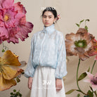 Woman in a light blue floral-patterned lace jacket and white skirt standing among large flowers with 'BWBYAZ' branding.