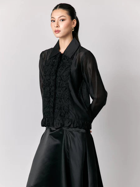 Woman wearing a black sheer shirt by Studio Tui with ruffled details on a plain background