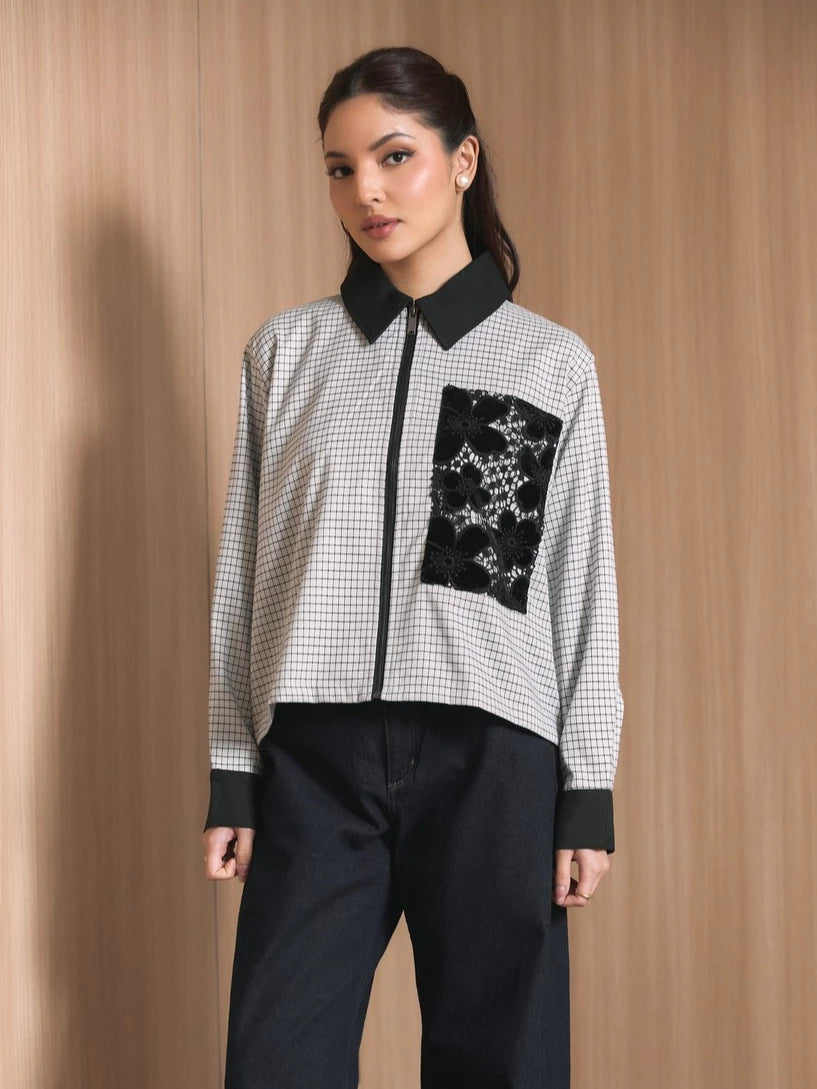 A woman modeling a long-sleeve zip-front jacket by Studio Tui with a black and white gingham pattern, solid black collar, cuffs, and hem, featuring a large black embroidered chest pocket. She is wearing dark wide-leg jeans against a light wood-paneled background.