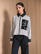 A woman modeling a long-sleeve zip-front jacket by Studio Tui with a black and white gingham pattern, solid black collar, cuffs, and hem, featuring a large black embroidered chest pocket. She is wearing dark wide-leg jeans against a light wood-paneled background.