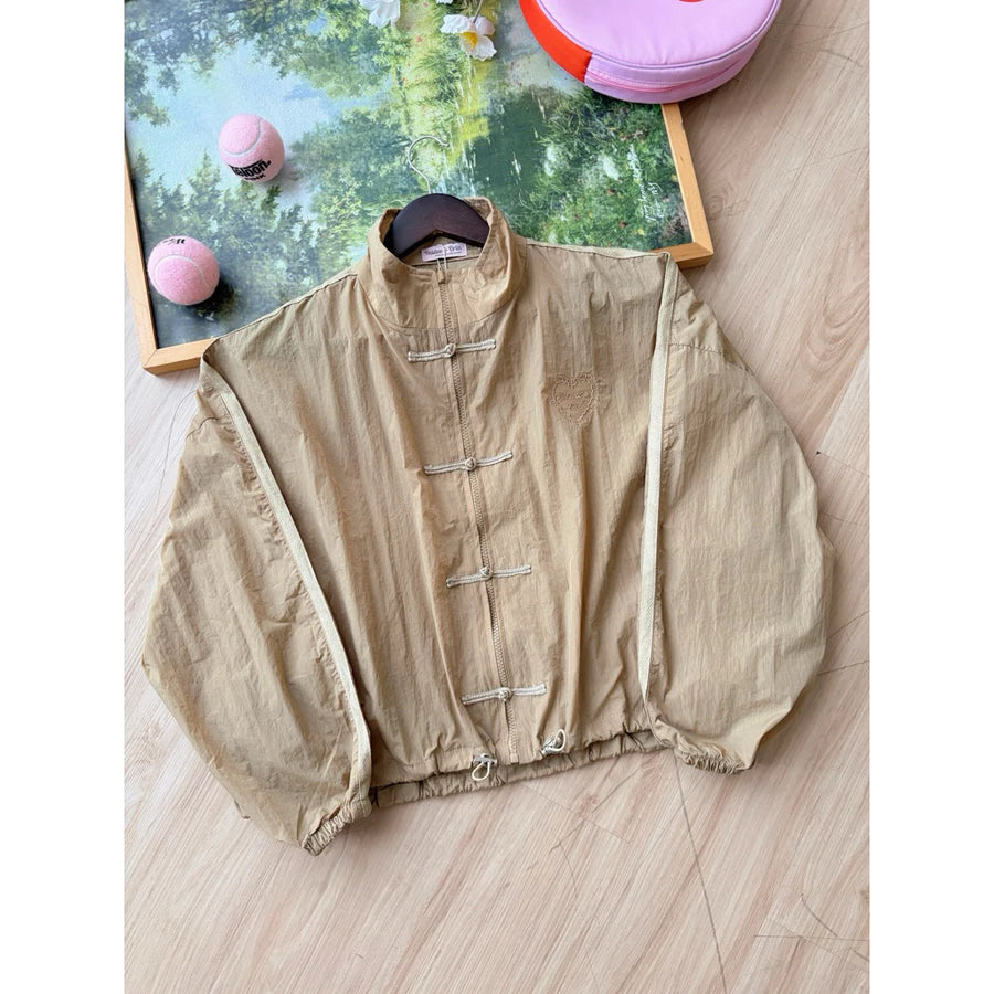 A beige cheongsam jacket by Haidee Orlin with a high collar, laid flat on a light wood floor with decorative props in the background.