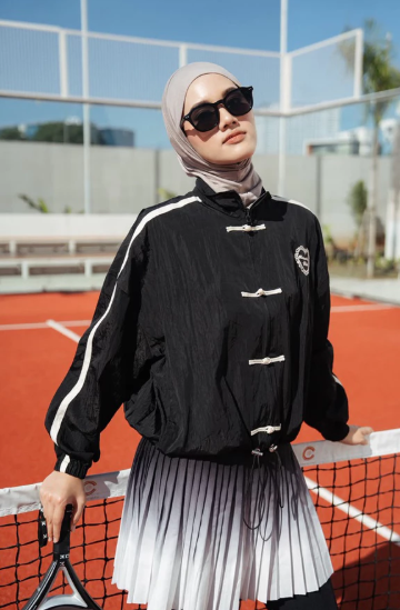 A woman wearing a black lightweight cheongsam jacket by Haidee Orlin with a black-and-white pleated skirt, posing on an outdoor tennis court.