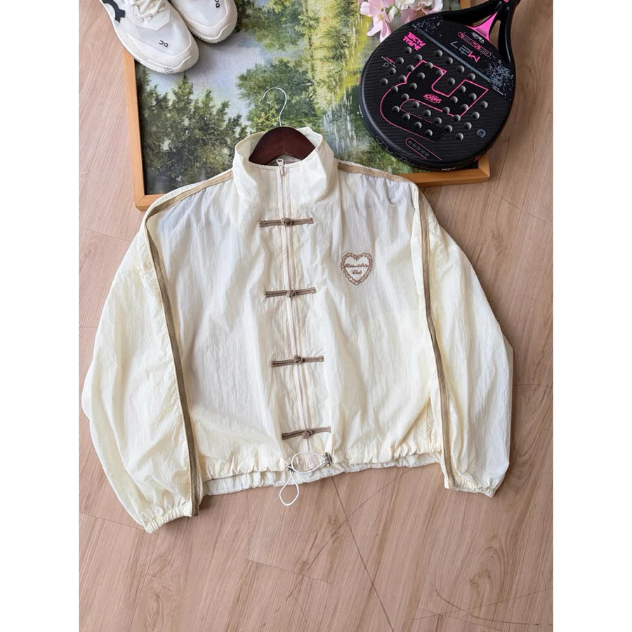 Cream-colored windbreaker cheongsam jacket by Haidee Orlin, laid flat on a wooden floor next to a framed picture, shoes, and a paddle racket.