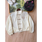 Cream-colored windbreaker cheongsam jacket by Haidee Orlin, laid flat on a wooden floor next to a framed picture, shoes, and a paddle racket.