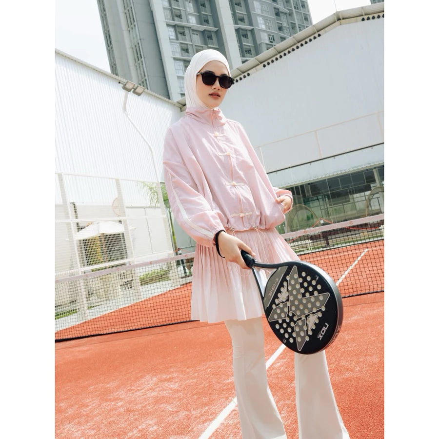 A woman wearing a light pink long-sleeve athletic cheongsam jacket by Haidee Orlin and white pants stands on an outdoor tennis court holding a black paddle racket.