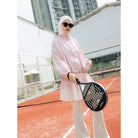 A woman wearing a light pink long-sleeve athletic cheongsam jacket by Haidee Orlin and white pants stands on an outdoor tennis court holding a black paddle racket.