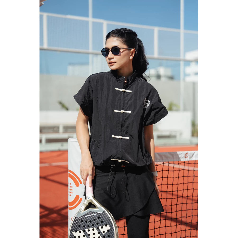 A woman wearing a black cheongsam active vest by Haidee Orlin with black pants stands on an outdoor tennis court holding a paddle racket.
