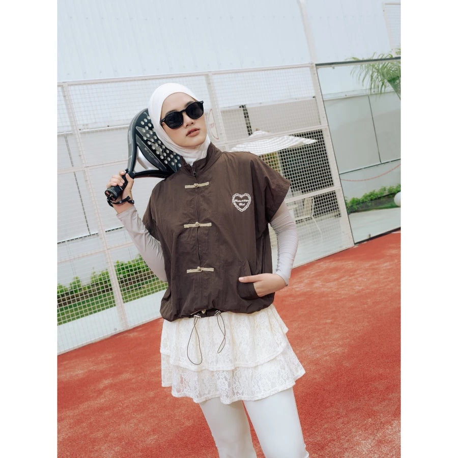 A woman wearing a brown cheongsam vest by Haidee Orlin over a white long-sleeve top and white lace skirt, posing on an outdoor tennis court while holding a racket.