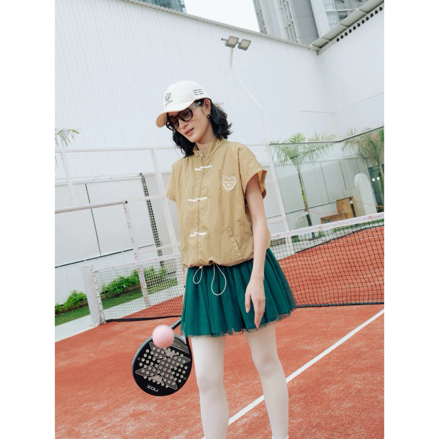 A woman stands on an outdoor tennis court wearing a khaki cheongsam vest by Haidee Orlin with white drawstrings, a dark green pleated tennis skirt, white tights, a white cap, and sunglasses. She holds a paddle with a pink ball.