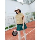 A woman stands on an outdoor tennis court wearing a khaki cheongsam vest by Haidee Orlin with white drawstrings, a dark green pleated tennis skirt, white tights, a white cap, and sunglasses. She holds a paddle with a pink ball.