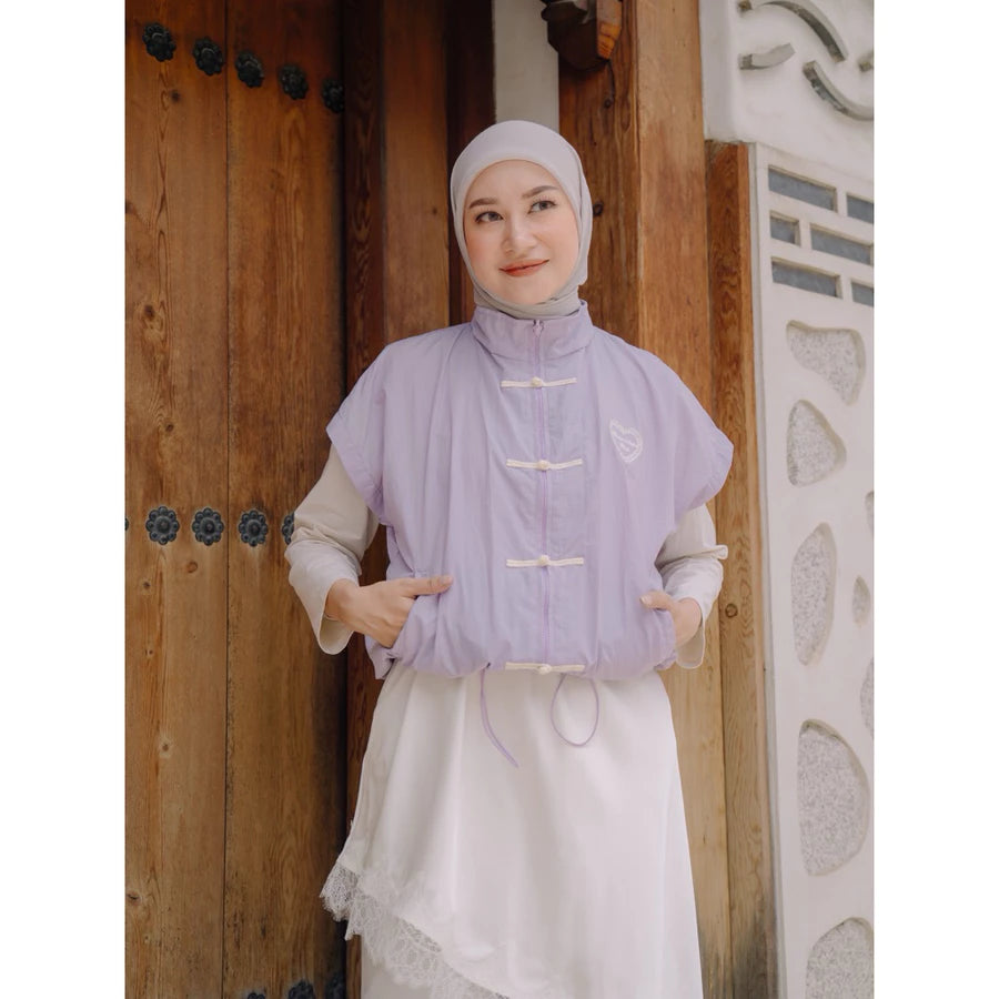A woman wearing a lilac sleeveless cheongsam vest by Haidee Orlin over a white dress and a white hijab, standing in front of a rustic wooden door.