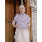 A woman wearing a lilac sleeveless cheongsam vest by Haidee Orlin over a white dress and a white hijab, standing in front of a rustic wooden door.