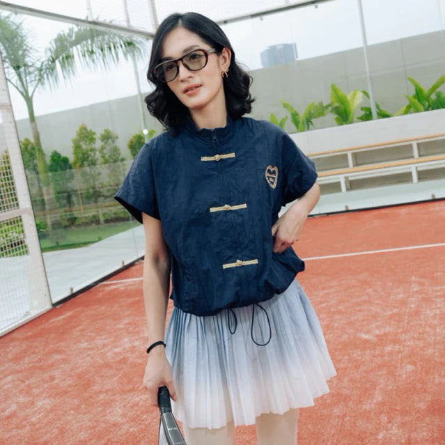 A woman wearing a navy blue cheongsam vest by Haidee Orlin with gold-toned frog closures and a light blue to white ombré tulle skirt, standing on an outdoor tennis court.