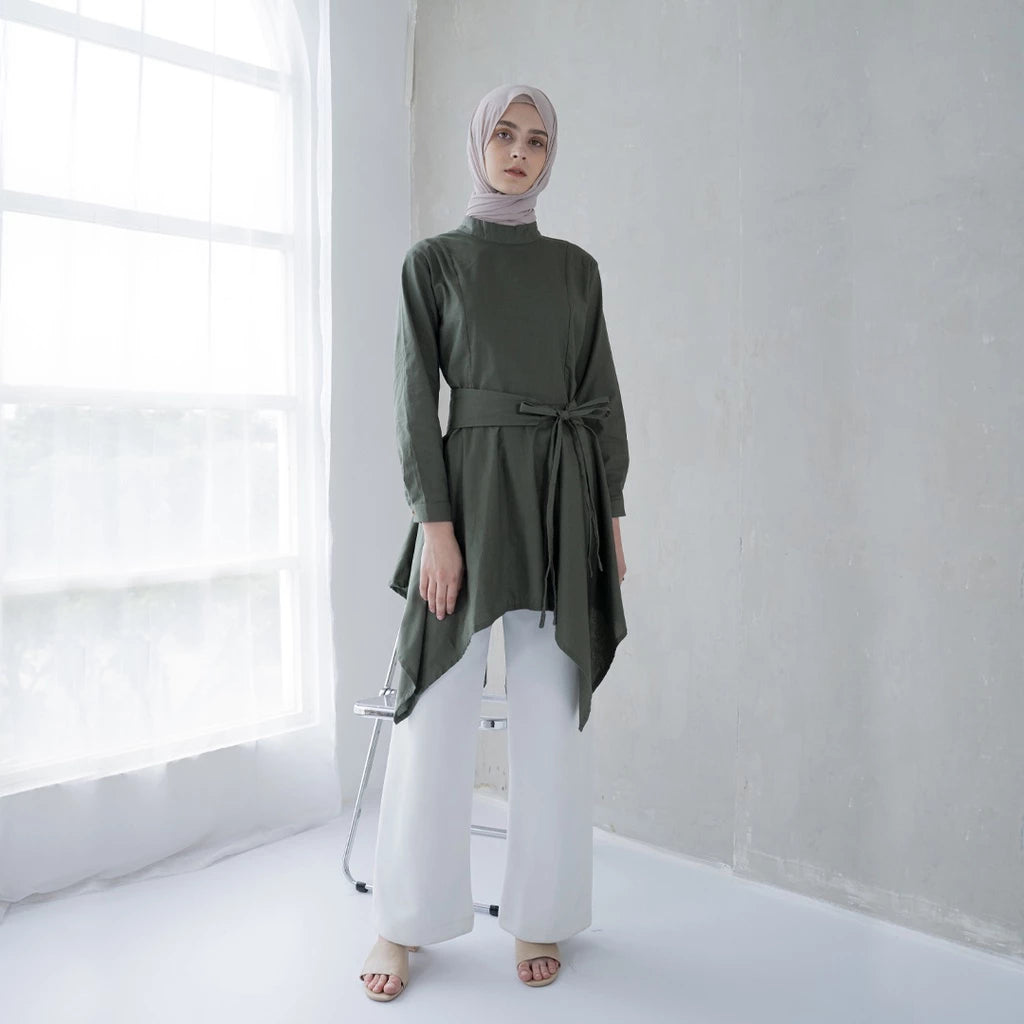 A woman wearing a green tunic with a semi turtleneck, sleeve pleats, and an detachable belt, paired with white pants.