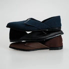 Three pairs of slip-on flat shoes stacked vertically on a light gray background. The top pair is dark navy blue denim with a black sole, the middle pair is black leather with a black sole, and the bottom pair is brown leather with a black sole.