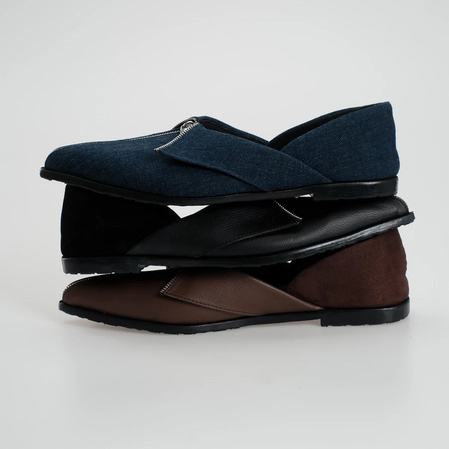 Three pairs of slip-on flat shoes stacked vertically on a light gray background. The top pair is dark navy blue denim with a black sole, the middle pair is black leather with a black sole, and the bottom pair is brown leather with a black sole.