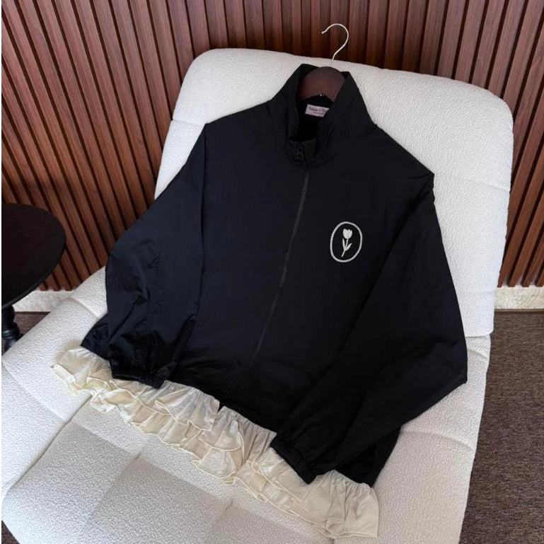 A black lightweight zip-up jacket by Haidee Orlin with a small white circular flower logo on the left chest, laid flat on a white textured chair in front of a brown slatted wall.