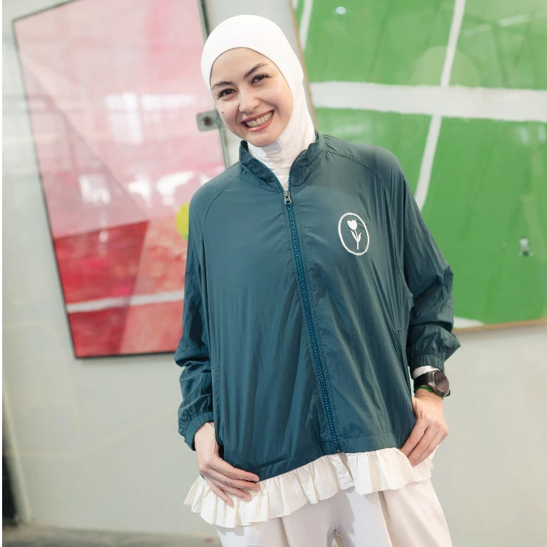 A woman wearing a dark teal green lightweight zip-up jacket by Haidee Orlin with a white ruffled hem, a white hijab, and white pants, standing in front of a modern wall with abstract art.