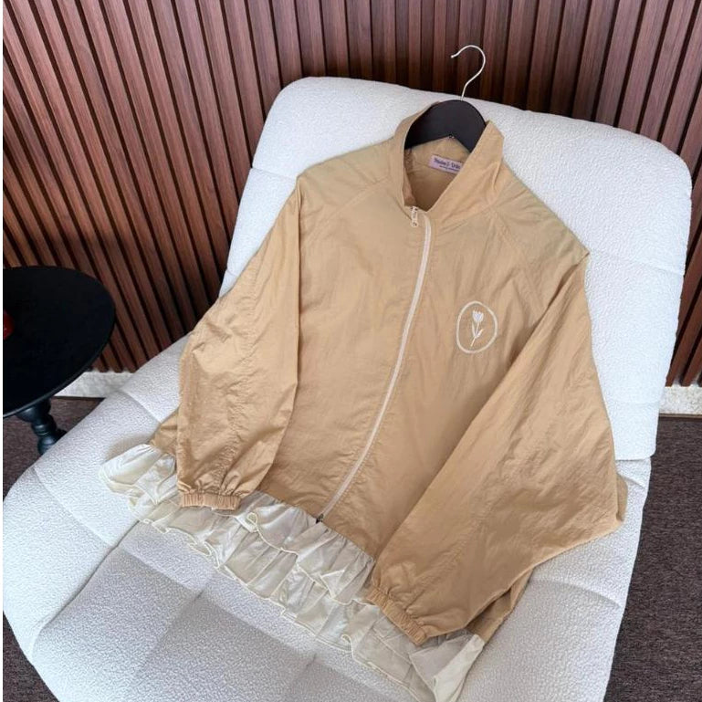 A beige lightweight zip-up jacket by Haidee Orlin with a small white circular logo on the chest, draped over a white textured chair in front of a brown slatted wall.